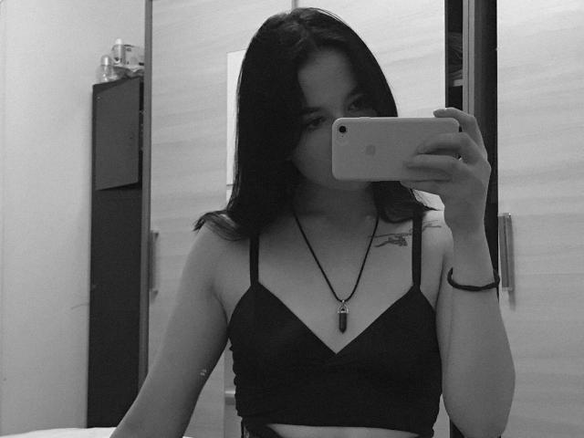 MertieDasinger taking a mirror selfie, wearing a black bralette, mask covering her mouth, holding a phone, necklace with a black pendant, tattoo on her right shoulder.