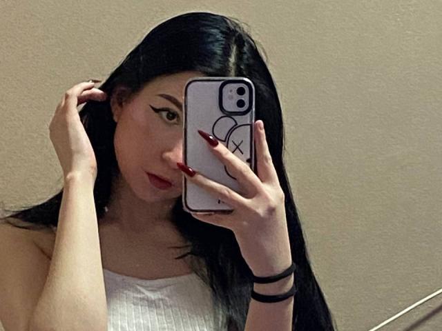 MertieDasinger with long black hair, red lipstick, white tank top, and red nails, holding a phone with an "X" case, taking a mirror selfie.