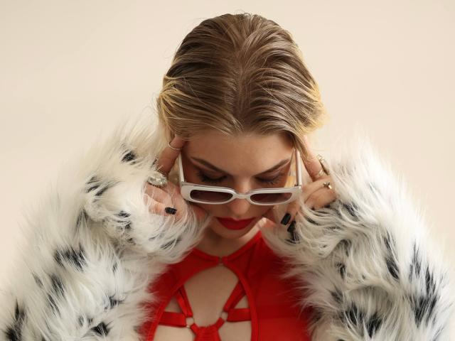 LesliNellenback with blonde hair, wearing white and black furry coat, red lingerie with strappy details, and white-framed glasses, touching ears, red lipstick, black nail polish.