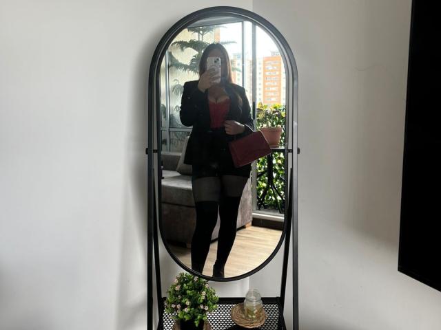 Gala Malkova steht vor einem Spiegel, trägt einen schwarzen Blazer, ein rotes Oberteil, schwarze Shorts und hohe Stiefel, hält eine rote Handtasche und hat ein Handy in der Hand..