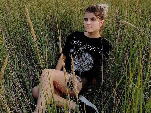 CoralieVonschriltz, with tied-up brown hair, sits in tall green grass wearing a black shirt with a dandelion and "NEVER DRY" text, crossed legs, serious expression.