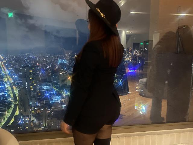 GalaMalkova with brown hair, black hat, black blazer, black mini-skirt, and black stockings, standing in front of a city skyline at night, showing her big ass and reflection.