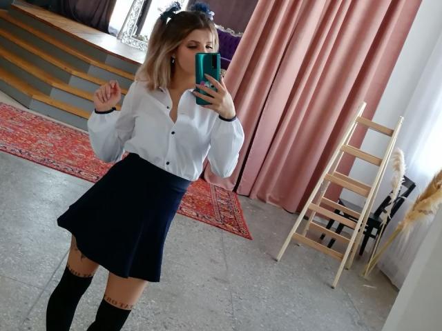 CoralieVonschriltz with blonde hair, white button-up shirt, dark blue skirt, black knee socks, holding green phone, standing in room with pink curtains, wooden ladder, and red rug.