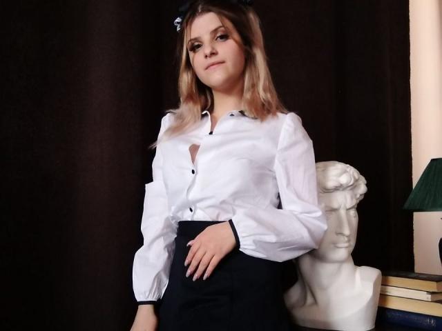 CoralieVonschriltz, white button-up shirt, black skirt, small tits, blonde hair, black bow, standing, hand on hip, white bust, books, green lamp, dark background.