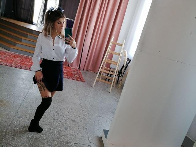 CoralieVonschriltz with blonde hair, white blouse, black skirt, knee socks, holding phone, mirror selfie, visible tattoo on thigh, standing in room with pink curtains and wooden stairs.