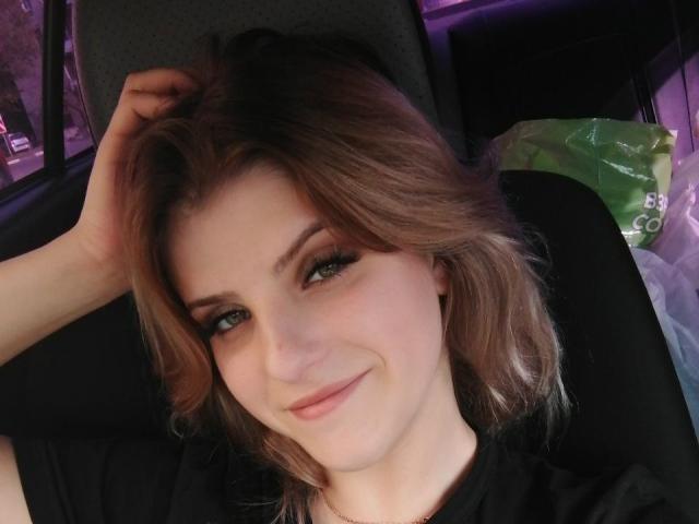 CoralieVonschriltz has light brown hair, green eyes, and a slight smile. She's lying in a car, hand in hair, wearing a black shirt, looking cute and relaxed.