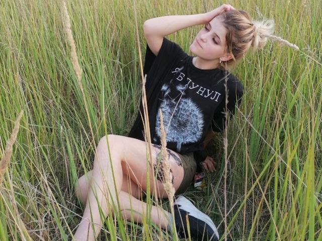 CoralieVonschriltz, blonde, sitting in tall grass, black t-shirt with white design, green shorts, hand in hair, legs crossed, relaxed, summer, casual, soft light, natural setting.