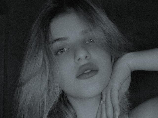 CoralieVonschriltz has pouty lips, light eyes, and messy blonde hair, resting her chin on her hand with a ring, in a grayscale, slightly blurred photo.