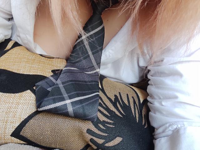 MinistraX's tits, covered by a white shirt, are partly exposed by a black and gray striped tie. Her pink hair is visible, and she's sitting on a patterned seat.