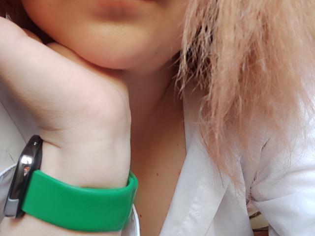MinistraX's pale hand rests on her chin, wearing a green wristband with a black metal clip, her pinkish-brown hair slightly messy, a white shirt partially visible.
