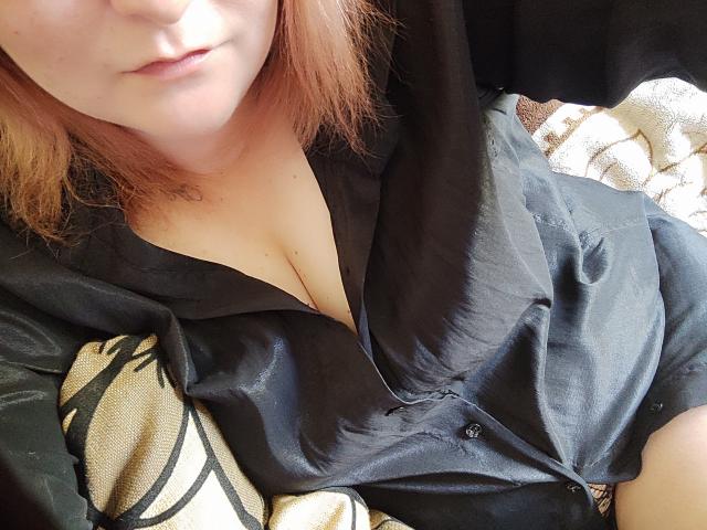 MinistraX with pale skin, pink hair, and slight smile, wearing a black shirt, showing cleavage, sitting on a beige and black patterned blanket, in a relaxed position.