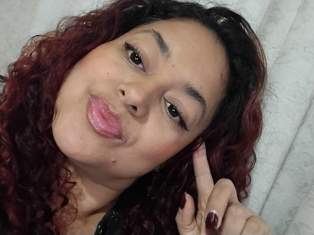 SophiLarusso with curly red-brown hair, light brown skin, and pink glossy lips, making a peace sign with her right hand, showing dark red nail polish.