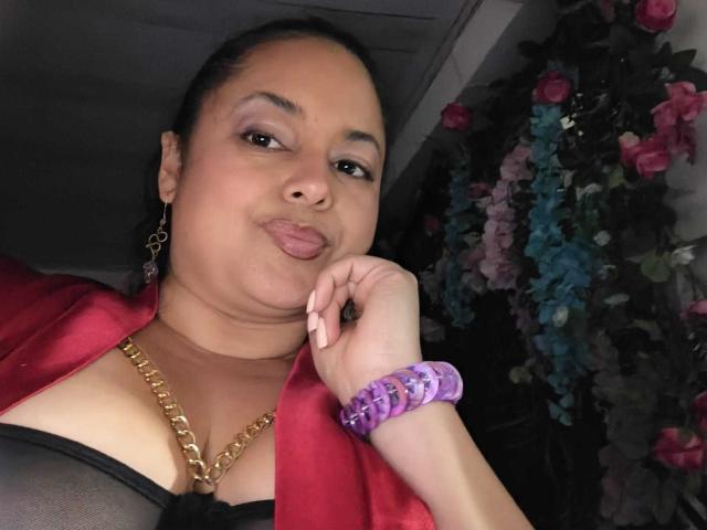 SophiLarusso with her puffy lips, gold chain, red satin robe, black mesh top, purple wristband, and gold earrings, lying down, looking at the camera, with a slight smile.