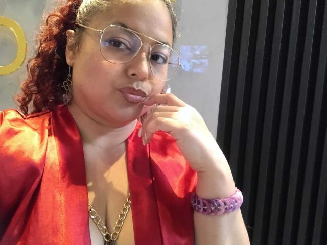 SophiLarusso with curly red hair, wearing glasses, a red satin robe, gold chain necklace, and purple bracelet, pouting with finger on chin, looking at camera.