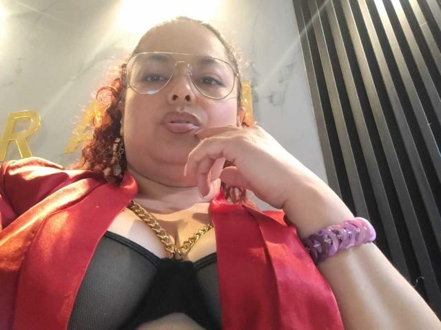 SophiLarusso with curly red hair, wearing clear glasses, red shirt, black bra, gold chain, purple bracelet, tongue out, hand near mouth, slightly chubby face, medium tits, light skin.