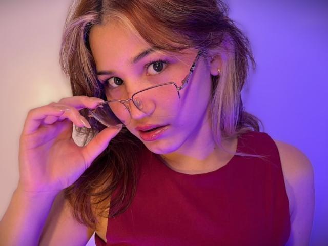 ElodieGardner with blonde hair, wearing a red sleeveless top, holding glasses in front of her face, purple-blue light on her skin, looking at the camera with parted lips.