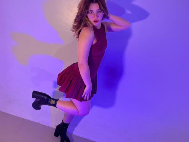 ElodieGardner with wavy brown hair, red dress, and black boots, standing against a wall with a purple-blue shadow, looking at the camera with a slightly open mouth.