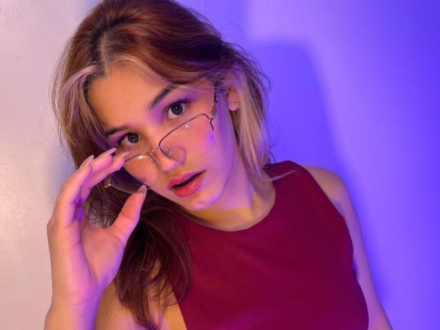 ElodieGardner with glasses, red top, and light brown hair, touching nose, looking surprised, blue and purple lighting, slightly open mouth, earrings, and a subtle smile.