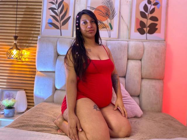Lunaarossee, une femme à la peau brune avec de longs cheveux noirs, portant une combinaison rouge, agenouillée sur le lit, montrant ses fesses, avec de petits tatouages sur la cuisse et le bras, regardant la caméra, chambre à coucher..