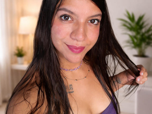 Lunaarossee, com cabelos longos e pretos, batom rosa e um colar roxo, segurando o cabelo, mostrando pequenas tatuagens e vestindo uma blusa roxa, sorrindo para a câmera..