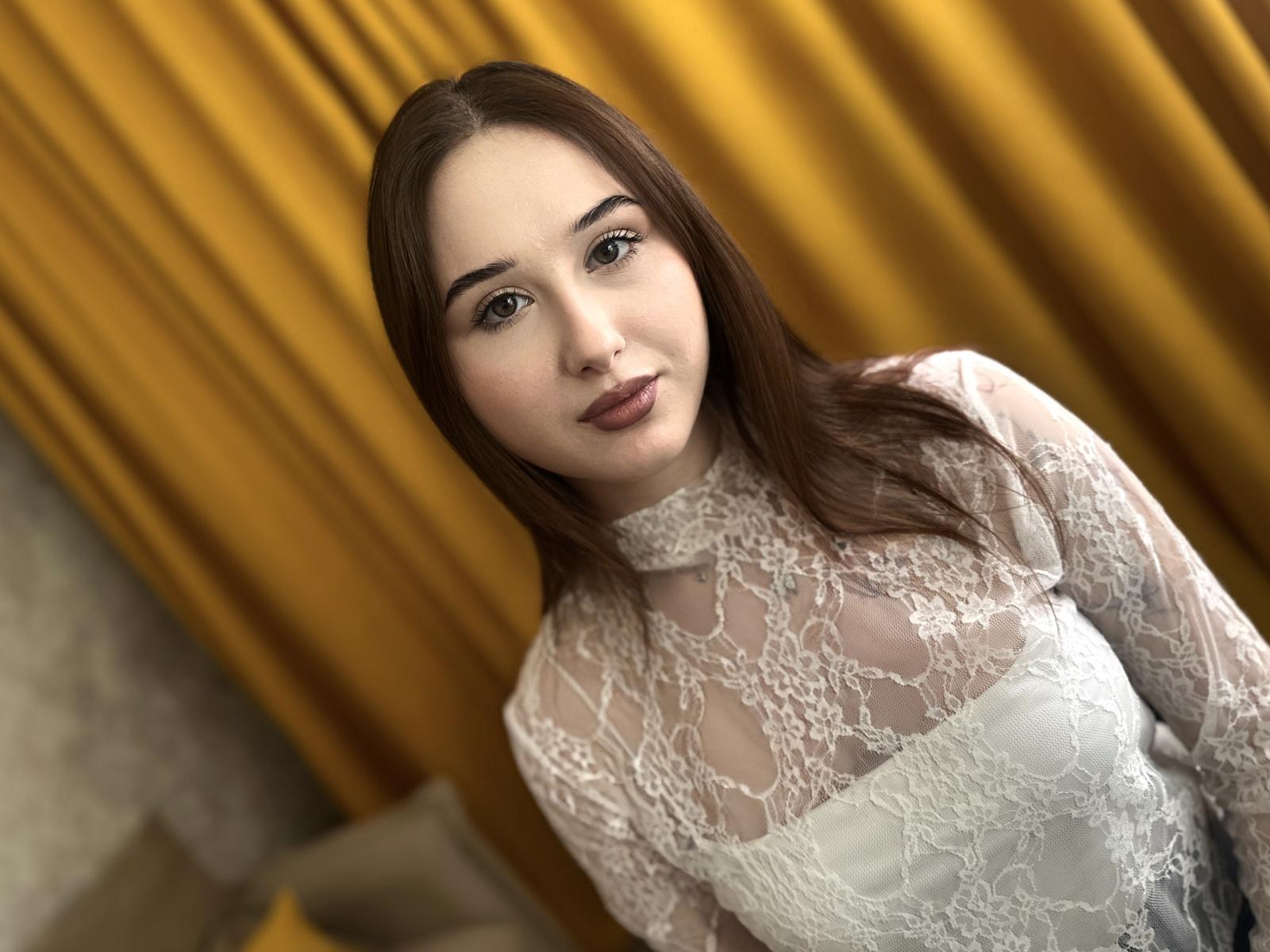 JulieVamp with brown hair, white lace top, showing her tits through sheer fabric, yellow curtain background, soft smile, subtle makeup, medium build, cute, and slightly tilted head.