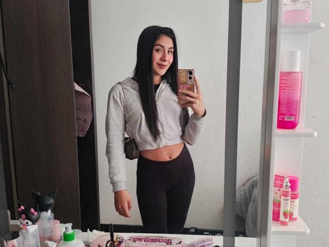 ChloeXk with long black hair, small tits, wearing a gray cropped hoodie and black pants, taking a mirror selfie in a bathroom, smiling, holding a pink phone.