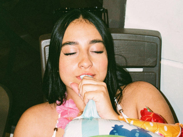 EmmaWaltter with black hair, eyes closed, sucking on a blue straw, wearing a colorful halter top, black sunglasses on head, sitting in a dark chair, slightly tan skin.