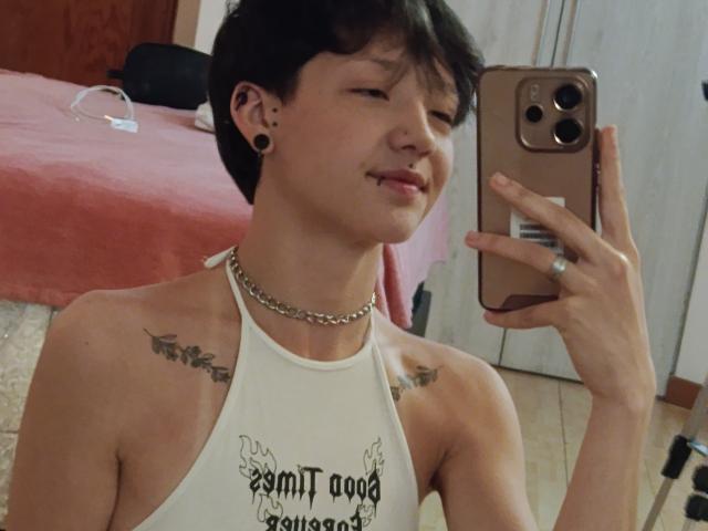 LissyLuxe with short black hair, small earrings, and a chain necklace, taking a mirror selfie with a smartphone, showing tattoos on shoulders, and wearing a white sleeveless top.