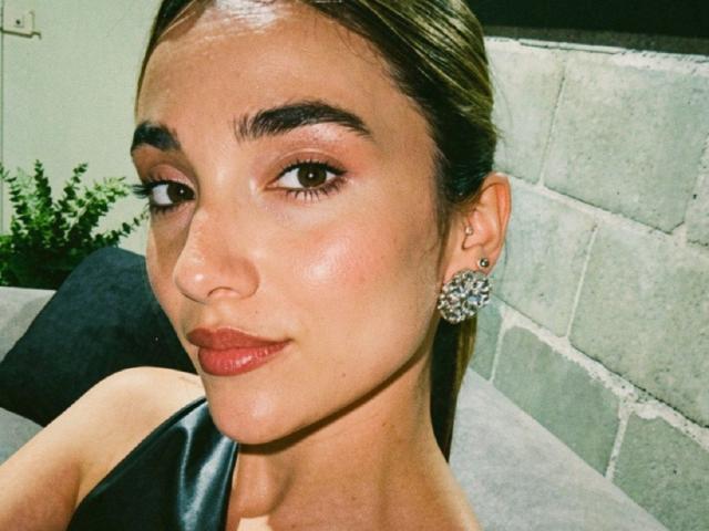AdharaCooperr with shiny skin, brown eyes, and full red lips, wearing a black top, and round, jeweled earrings, has brown hair pulled back. Her tits are not visible.