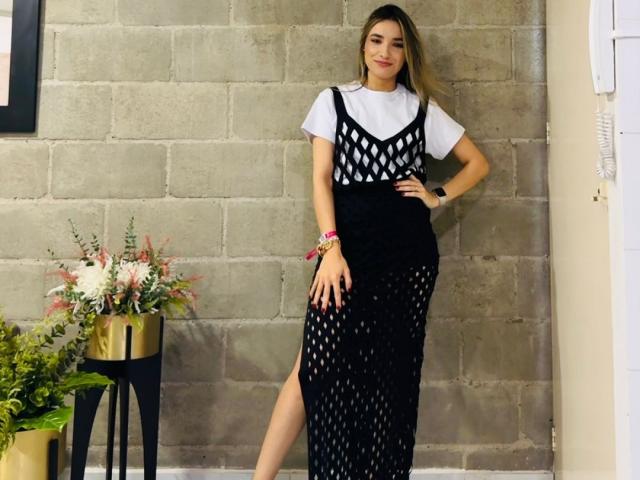 AdharaCooperr steht in einem schwarzen Netzkleid mit weißem Oberteil, eine Hand am Hüfte, blonde Haare, lächelnd, trägt ein rosa Armband, eine schwarze Uhr, schwarze Leggings und ein weißes Hemd..