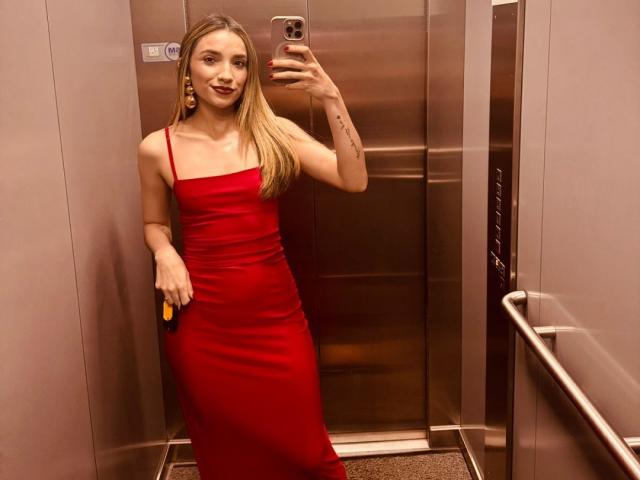 AdharaCooperr with long blonde hair, wearing a red dress, holding a phone, taking a mirror selfie in an elevator, showing her tits and a tattoo on her arm.