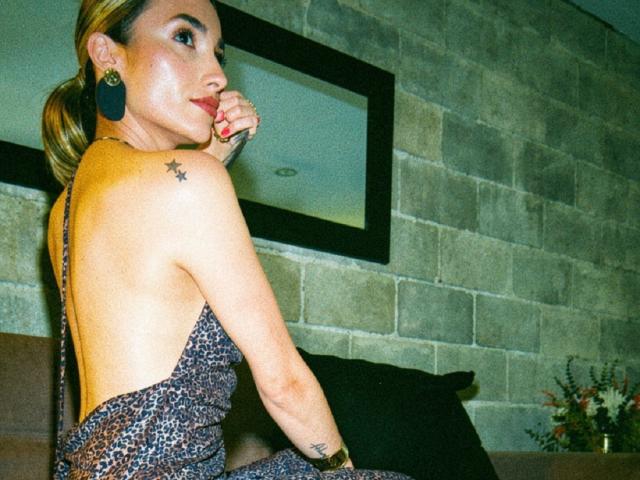 AdharaCooperr with back to camera, leopard-print dress, bare back, star tattoo on right shoulder, gold earrings, red nail polish, sitting on brown couch, looking over shoulder.