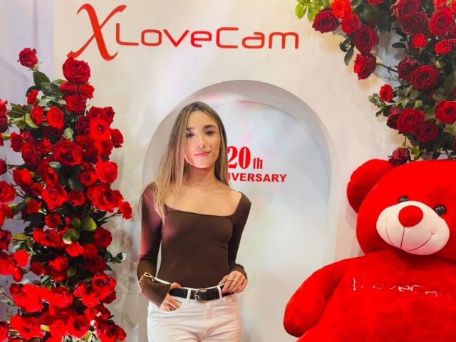 AdharaCooperr with long blonde hair, brown off-shoulder top, white pants, standing in front of red roses, giant red teddy bear, smiling, hands in pockets.
