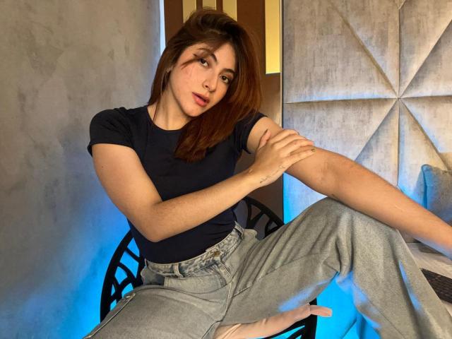 SpencerLe with brown hair, wearing a black top and gray pants, sitting with one leg up, holding her arm, showing a small tattoo on her wrist.