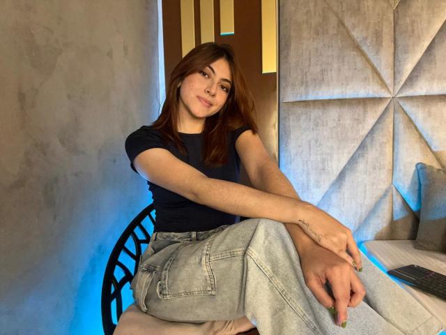 SpencerLe sits with legs crossed, wearing a black t-shirt and loose jeans, arms resting on knees, brown hair, subtle smile, tattoo on right wrist, in a modern room.