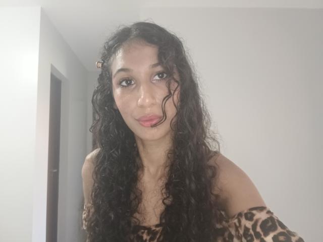AngelRosa69 has curly black hair, brown eyes, and is wearing a leopard print off-shoulder top, looking at the camera with a slight smile, her tits visible.