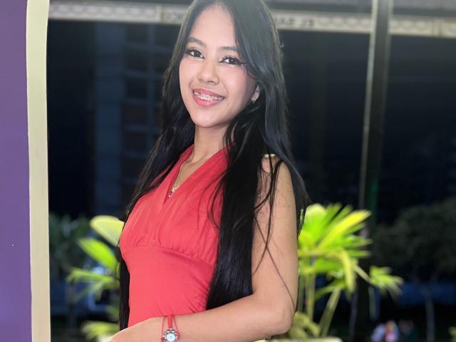 EstrellaXHot with long black hair, red sleeveless top, smiling, pink lipstick, silver watch, green plants in background, nighttime, purple wall on left.