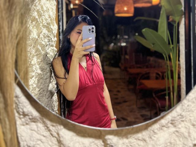 EstrellaXHot with long black hair, wearing a red halter dress, taking a selfie in a mirror. Her tits are covered by the dress. Her phone has a white case.