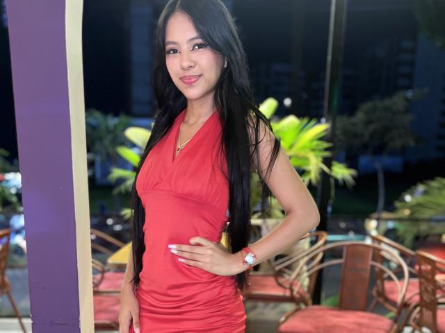 EstrellaXHot with long black hair, red dress, small tits, and hand on hip, smiling, wearing a red wristwatch, standing in a brightly lit outdoor area.