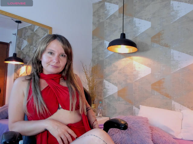 SharoLoveDion in a red dress with cutout, showing her small tits, sitting in a chair, smiling, blonde hair, arm tattoo, black lamp, purple pillow, beige wall.