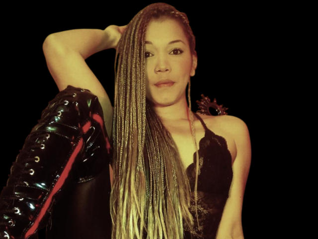 EmperatrizFetishist with long braided hair, black lace top, arm raised, holding shiny black leather with red lining, black background, confident expression, medium shot.