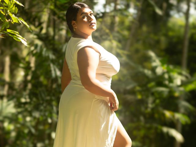 DojaAissha with big tits, curvy ass, and a thick, sexy body, wearing a white dress, standing in sunlight with green foliage, looking up, her dark hair in a bun.