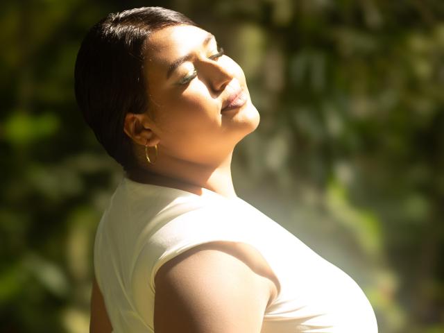 DojaAissha with short, slicked-back black hair, eyes closed, and head tilted up, wearing a white top, sunlight on her tan skin, gold hoop earrings, and a relaxed, happy expression.