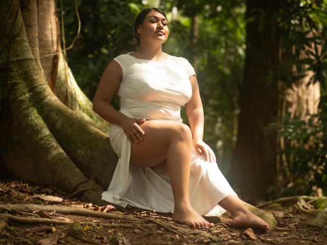 DojaAissha with big boobs, thick ass, and a curvy body sits on forest ground, wearing a white dress, barefoot, hands on knees, sunlight through trees, looking up.