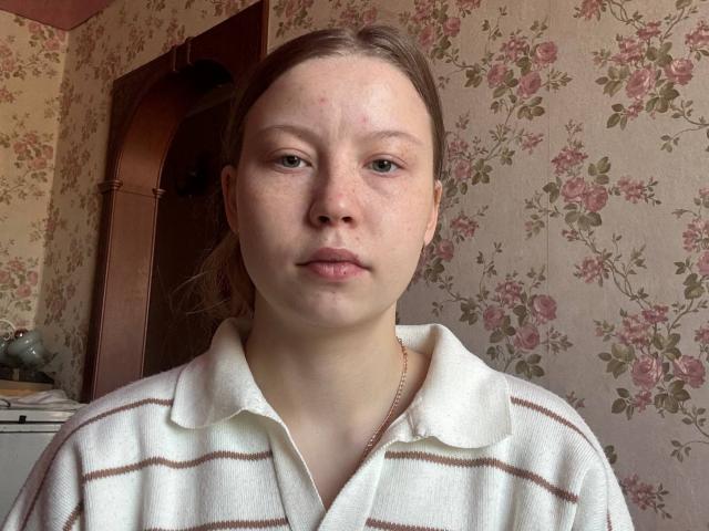 LoretaFeltner has a pale, freckled face, light brown hair in a ponytail, wearing a white, striped polo shirt, looking straight at the camera with a neutral expression.