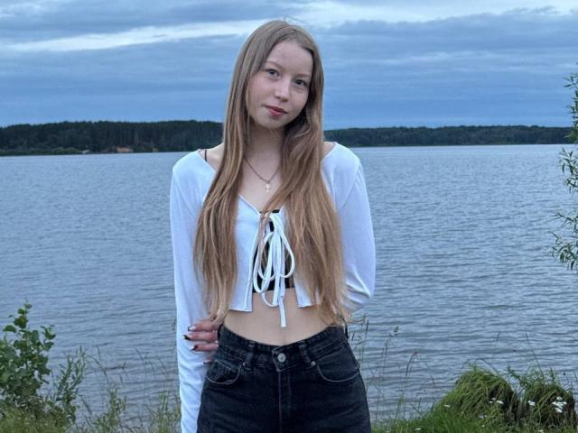 LoretaFeltner has long blonde hair, small tits, wearing a white long-sleeve crop top with white lace and black high-waisted jeans, standing by a lake.