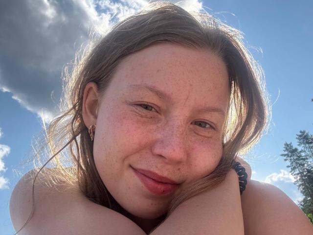 LoretaFeltner's face, freckled with light brown hair, smiling slightly, hands near her chin, blue sky and clouds, small earrings, black bracelet, sunlit, bare shoulders.