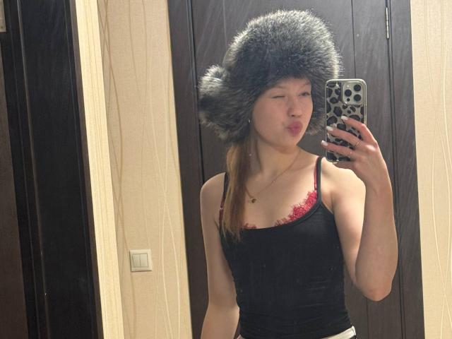 LoretaFeltner, wearing a grey furry hat, black tank top with red lace trim, pouting, taking a mirror selfie with a phone in a dimly lit room.