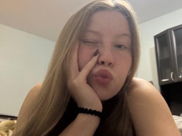 LoretaFeltner with blonde hair, winking, pouting lips, hand on cheek, black bracelet, wearing a sleeveless top, in a bedroom. Her tits are partially visible.