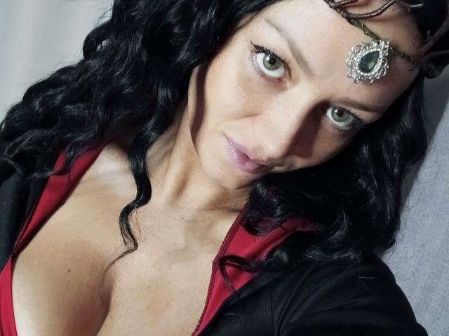 EhidnaLowq mit welligem, schwarzem Haar, grünen Augen und einem mit Juwelen besetzten Kopfschmuck, trägt ein rotes und schwarzes Oberteil, das ihre großen Brüste zeigt, und hat ein leichtes Lächeln..
